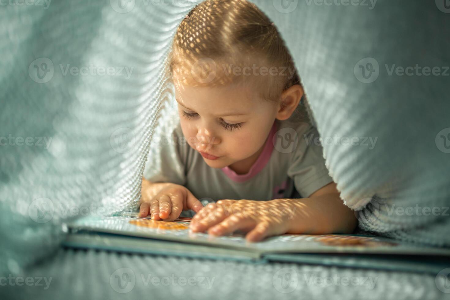 Little girl enjoying of review a book under blue knitted plaid in sunny morning photo