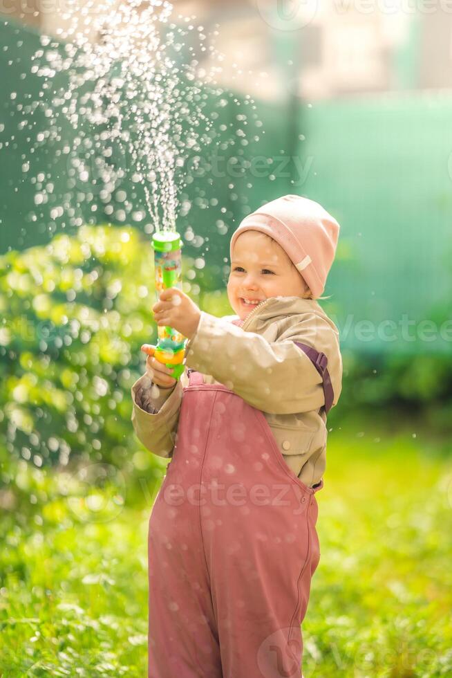 contento riendo niño niña 2-3 año antiguo vistiendo impermeable ropa tener un divertido con agua pistola en hogar patio interior foto