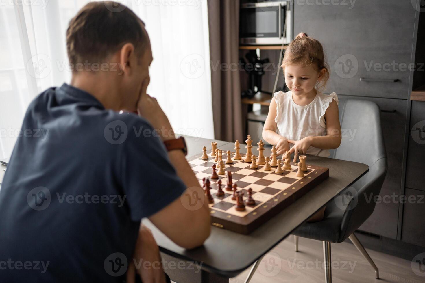 padre enseñando su pequeño hija a jugar ajedrez a el mesa en hogar cocina. el concepto temprano infancia desarrollo y educación. familia ocio, comunicación y recreación. foto