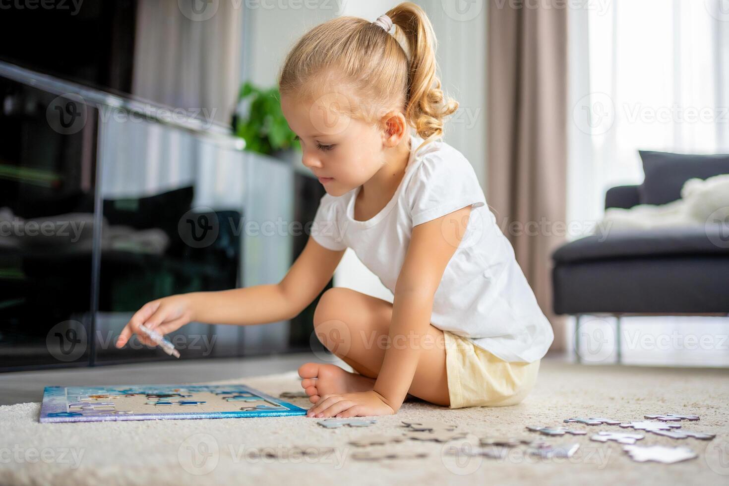 Little blonde girl sits at home on the carpet and collects puzzles photo