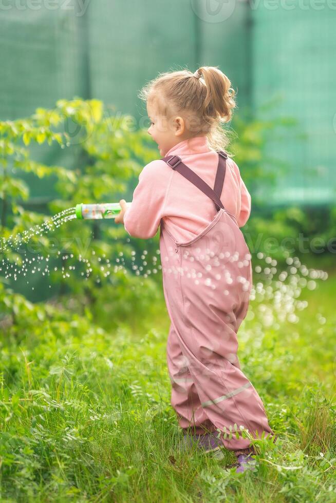 contento riendo niño niña 2-3 año antiguo vistiendo impermeable ropa tener un divertido con agua pistola en hogar patio interior foto