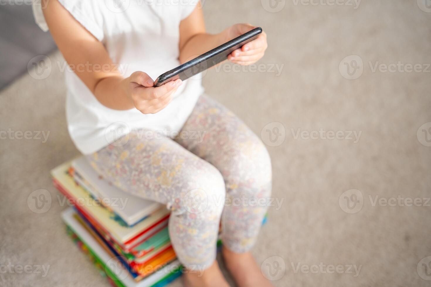 Child sits on a stack of children's fairy-tale books and watches cartoons on smartphone photo