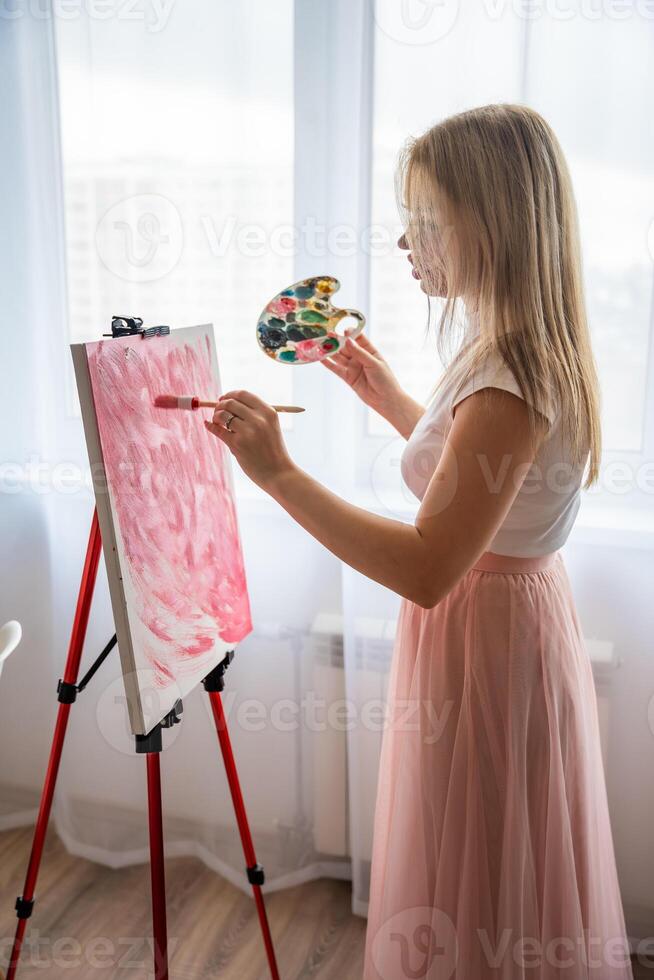 Young woman artist with palette and brush painting abstract pink picture on canvas near window. Art and creativity concept. High quality photo