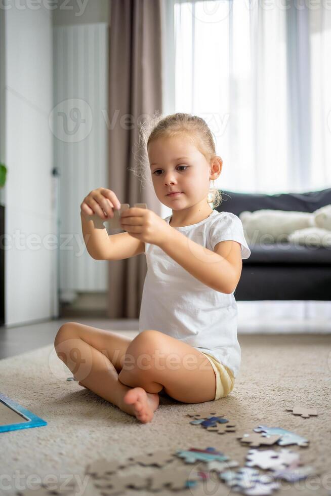 Little blonde girl sits at home on the carpet and collects puzzles photo