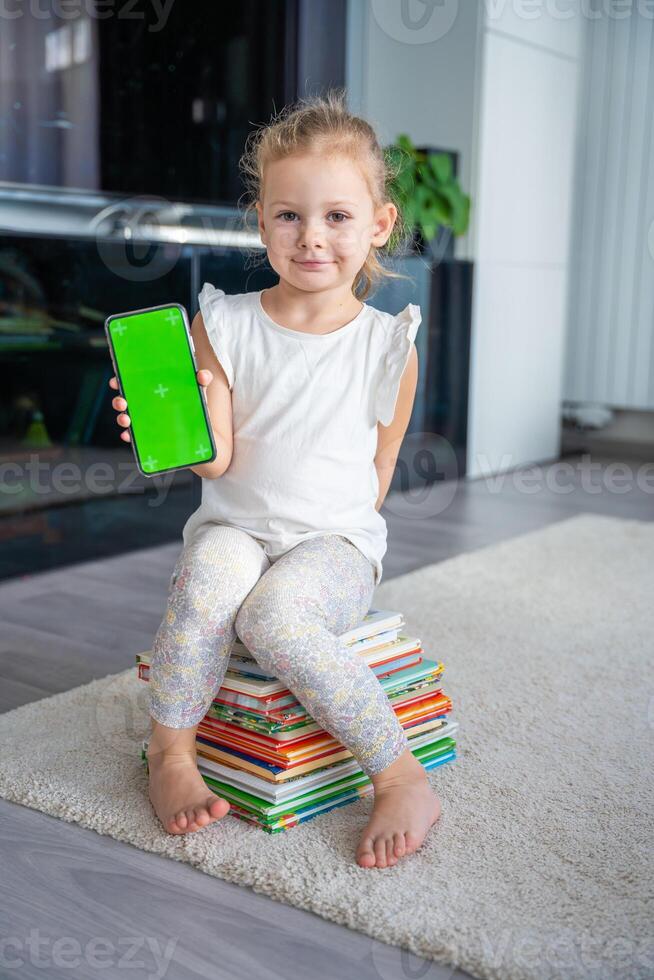 Little girl is sitting on stack of children's books and holding a smartphone with a chromakey photo