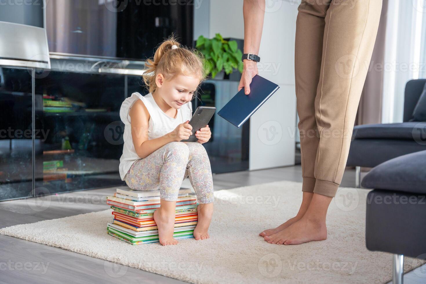 pequeño niña se sienta en apilar de para niños libros y usos su teléfono inteligente mientras madre da su un libro foto