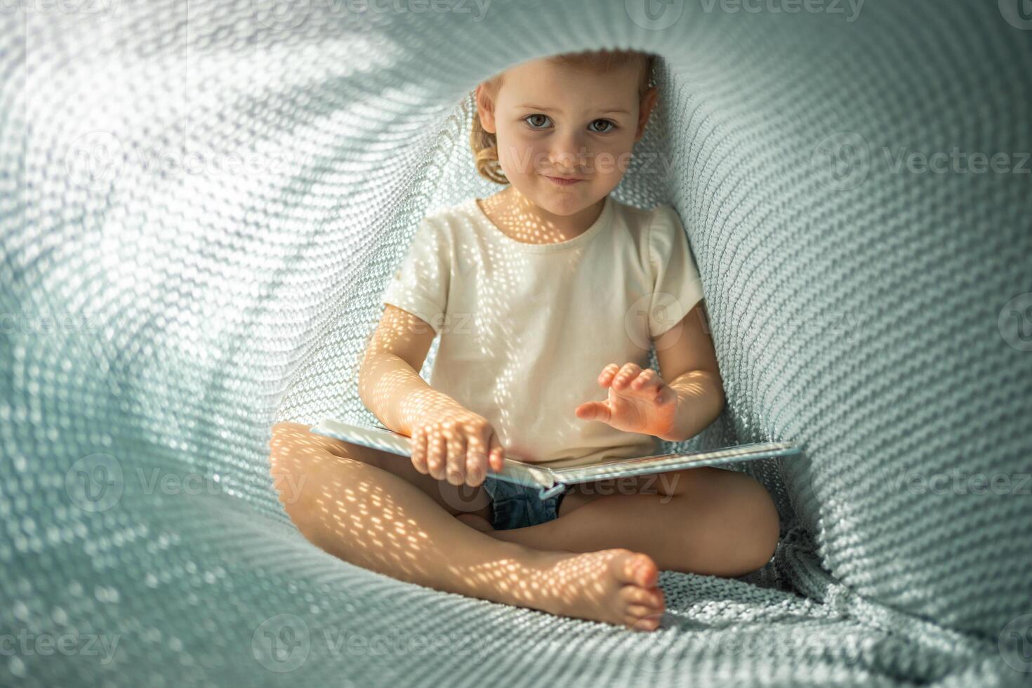 pequeño niña disfrutando de revisión un libro debajo azul de punto tartán en soleado Mañana foto