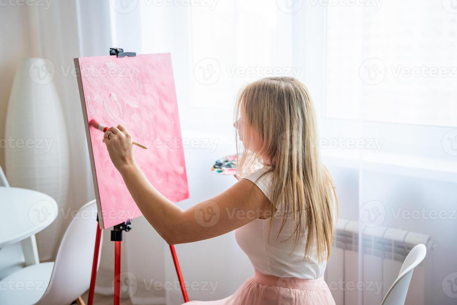 Young woman artist with palette and brush sitting and painting abstract pink picture on canvas near window at home. Art and creativity concept. High quality photo