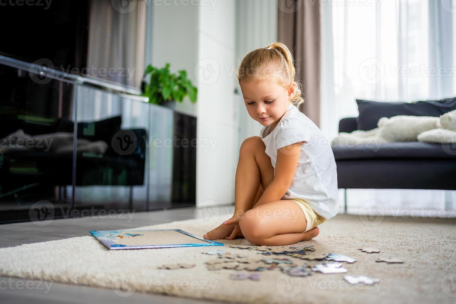 Little blonde girl sits at home on the carpet and collects puzzles photo