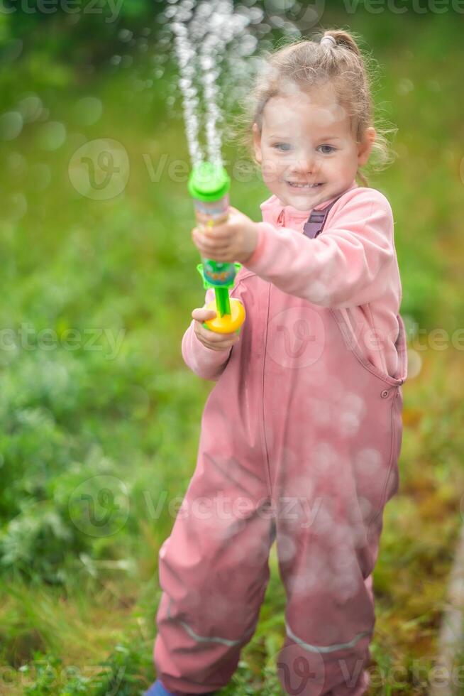 contento riendo niño niña 2-3 año antiguo vistiendo impermeable ropa tener un divertido con agua pistola en hogar patio interior foto