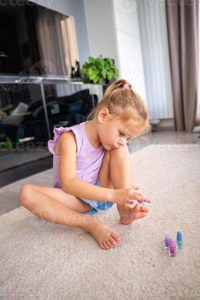 pequeño niña haciendo pedicure y pintura uñas con vistoso rosa, azul y púrpura uña polaco a hogar vivo habitación foto