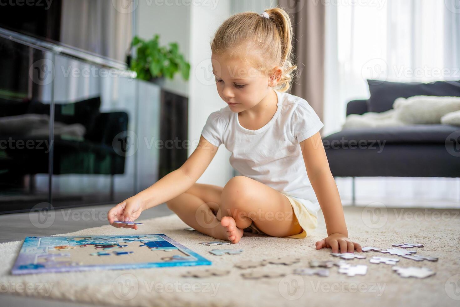 Little blonde girl sits at home on the carpet and collects puzzles photo