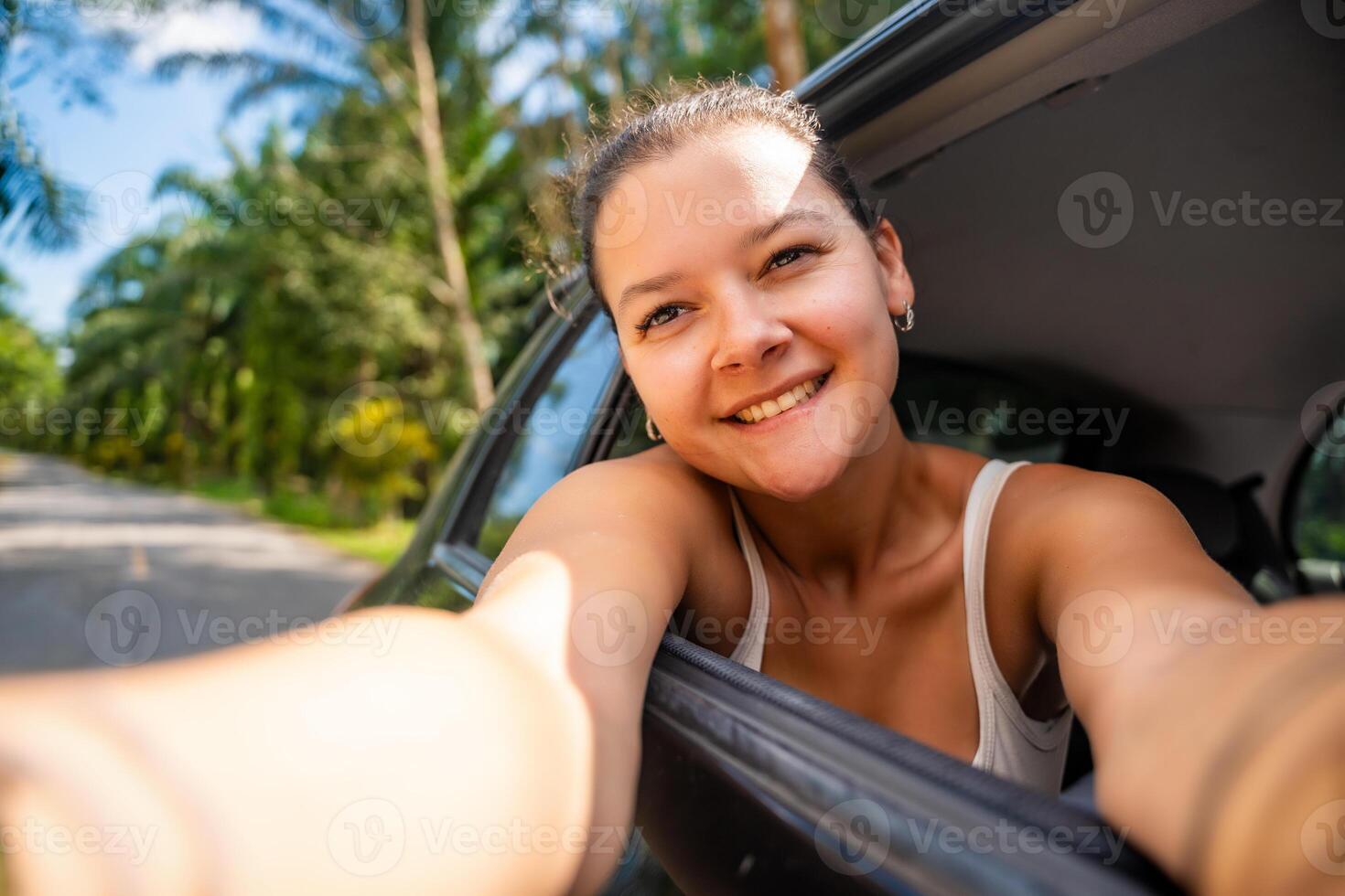Young woman with dark hair stuck her hands out of the cab of a car and make selfie on the road, concept of auto travel. High quality photo
