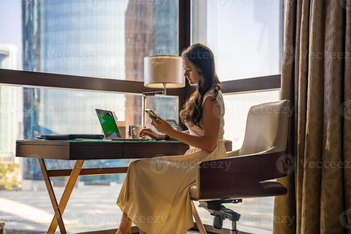 Stylish businesswoman freelancer working with laptop and smartphone, she is sitting on chair, enjoying panoramic view in city background. Low key photo. High quality photo