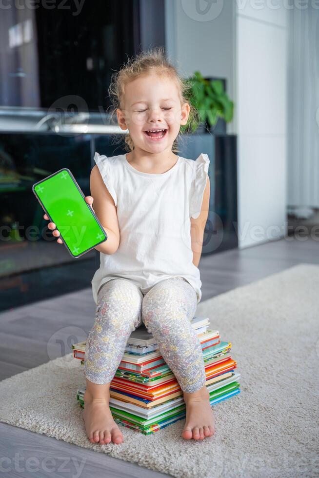 Happy Little girl is sitting on stack of children's books and holding a smartphone with a chromakey photo