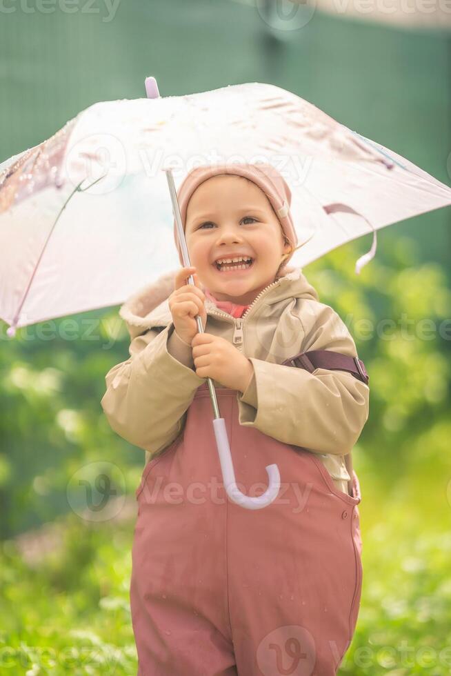 Happy laughing child girl 2-3 year old wearing waterproof clothes and holding pink umbrella have a fun on home backyard in rainy day photo