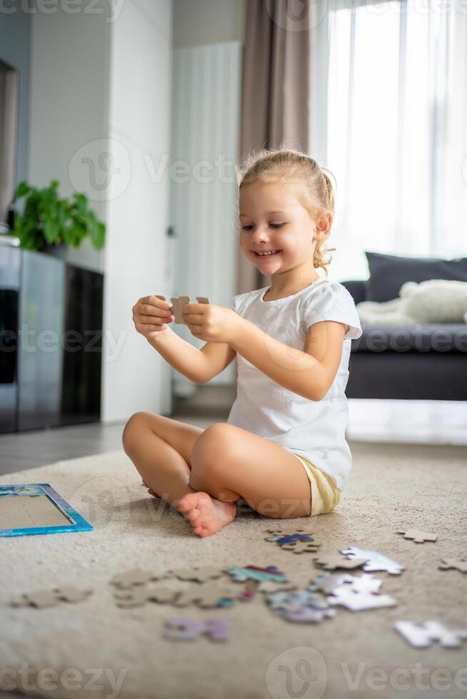 Little blonde girl sits at home on the carpet and collects puzzles photo