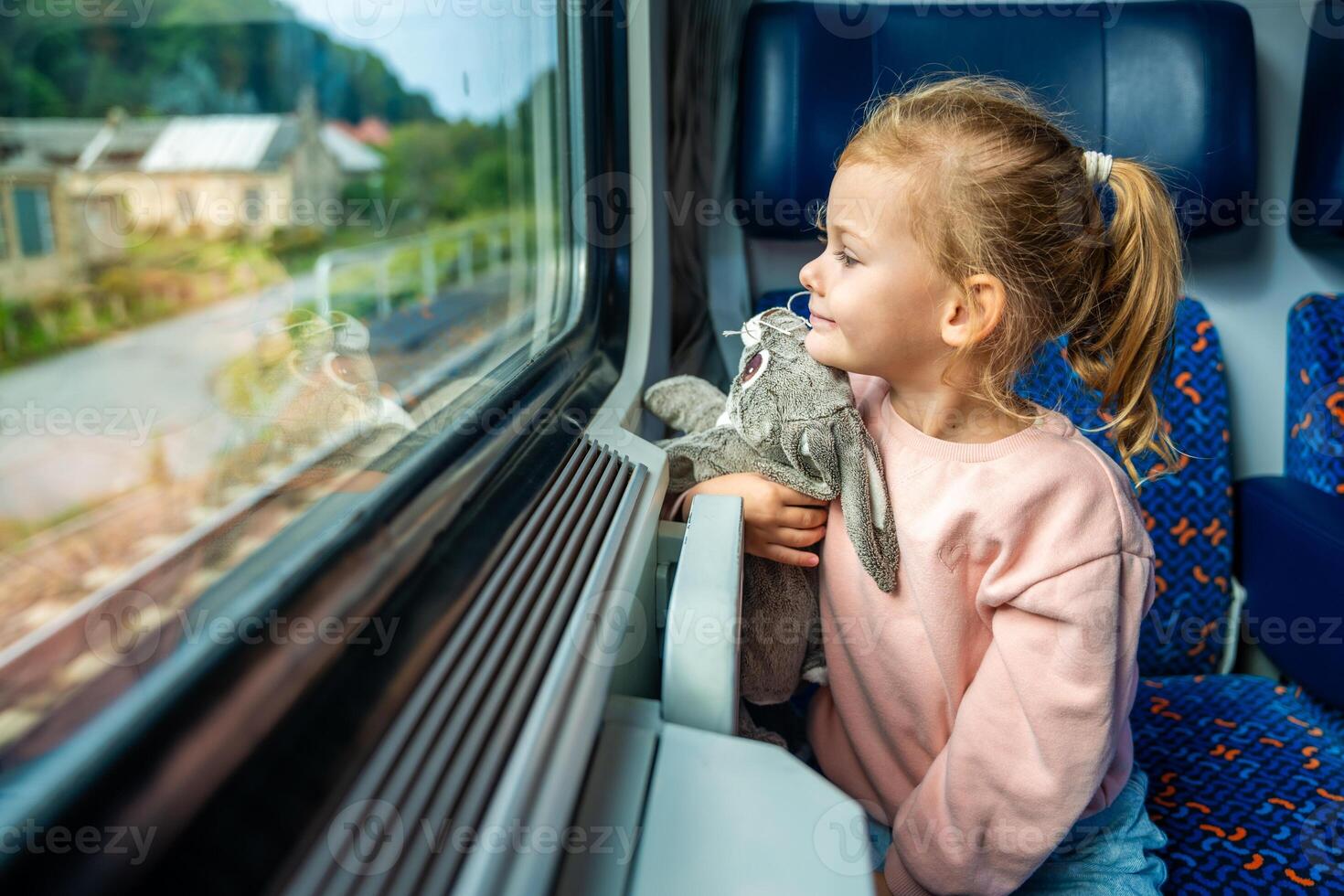 Smiling little girl with toy looking out train window outside, while it moving. Traveling by railway, Europe photo