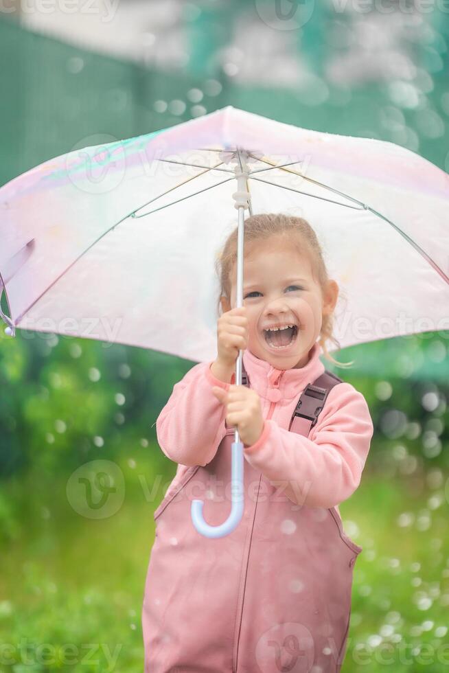 Happy laughing child girl 2-3 year old wearing waterproof clothes and holding pink umbrella have a fun on home backyard in rainy day photo