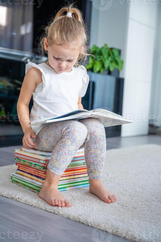 Little girl is sitting on stack of children's books and leafing through a book with fairy tales photo