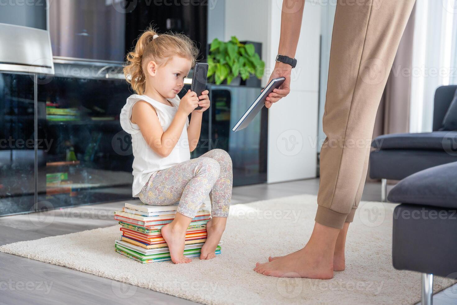 Little girl sits on a stack of children's books and uses her smartphone while mother gives her a book photo