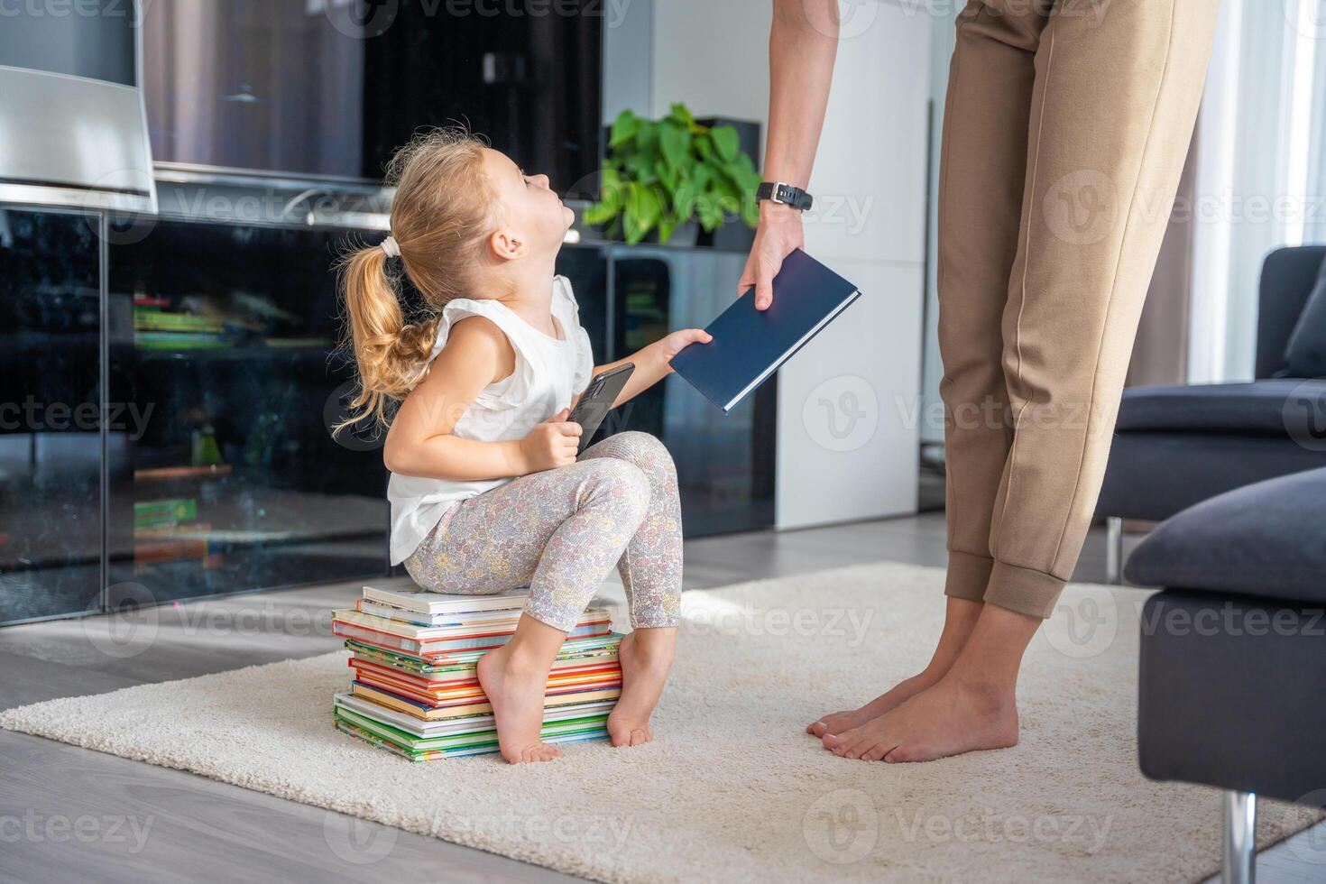 Little girl sits on a stack of children's books with smartphone while mother gives her a book photo