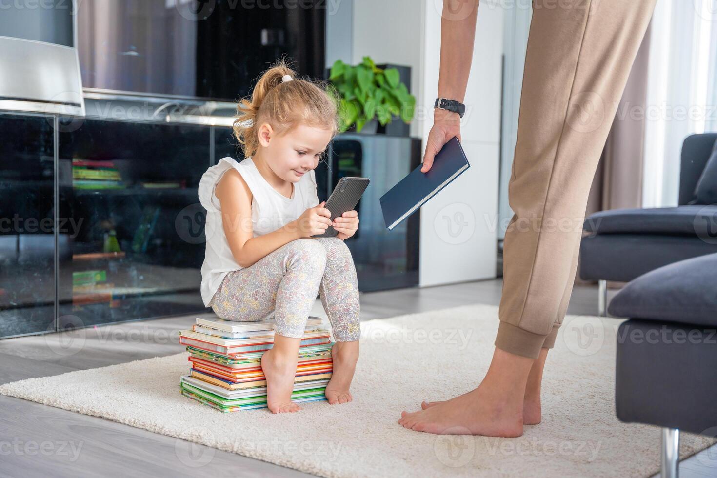 Little girl sits on a stack of children's books and plays with her smartphone while mother gives her a book photo