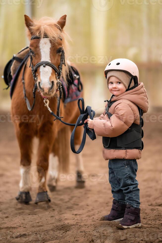 Portrait of little girl in protective jacket and helmet with her brown pony before riding Lesson. High quality photo