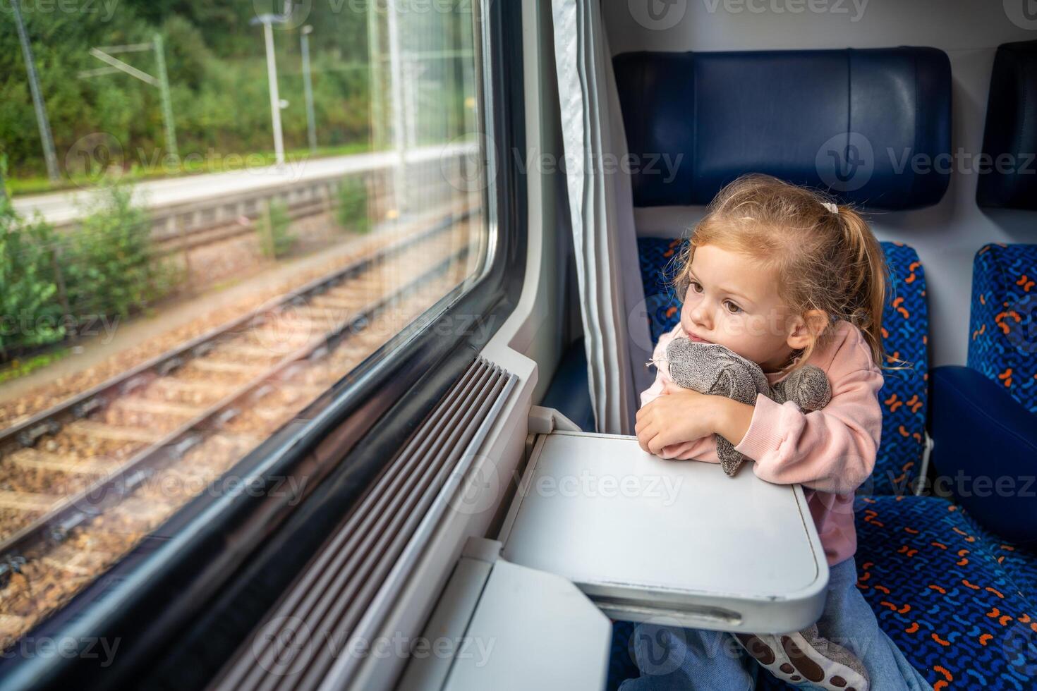 Bored little girl with toy looking out train window outside, while it moving. Traveling by railway, Europe photo