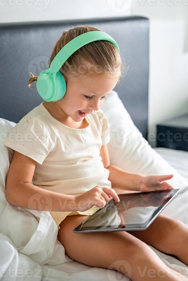 Little girl in headphones using digital tablet and smiling happy while listening to music or playing game in home bed. High quality photo