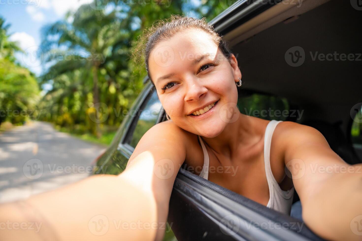 Happy young woman with dark hair stuck her hands out of the cab of a car and make selfie on the road, concept of auto travel. High quality photo