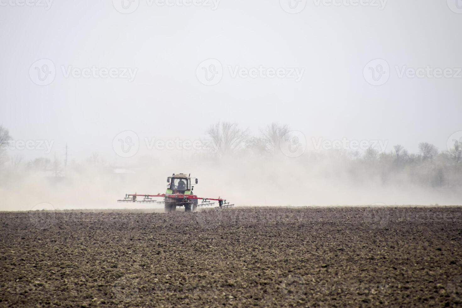 The tractor harrows the soil on the field and creates a cloud of dust behind it photo