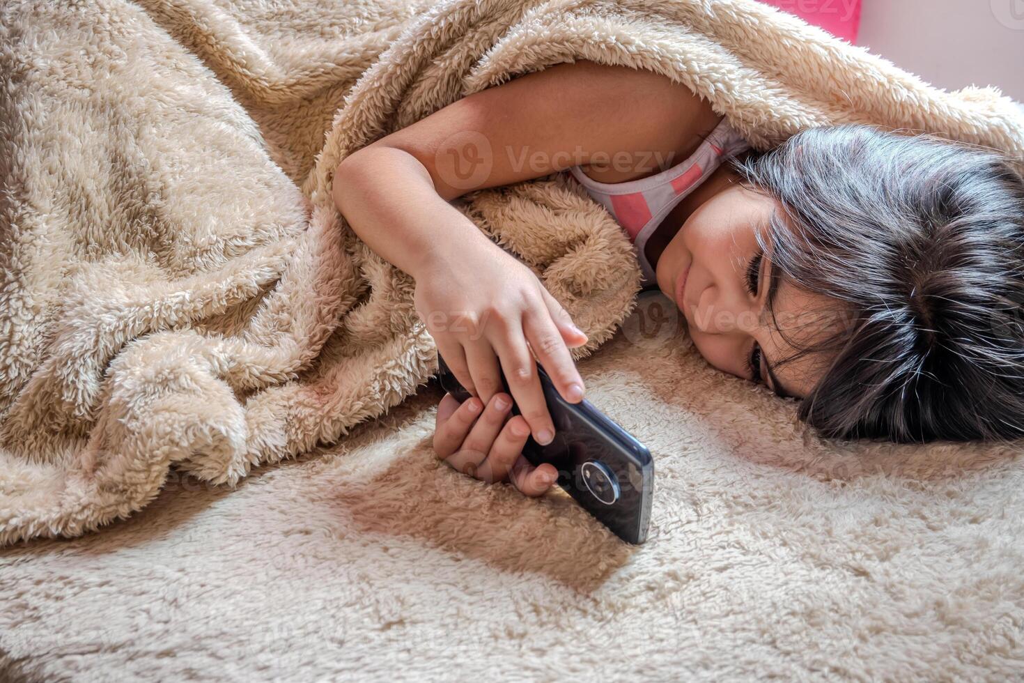 linda pequeño niña utilizando un teléfono inteligente niño jugando con móvil teléfono, acostado en un cama. tiempo libre. tecnología y Internet concepto. niñito tendido en cama y mirando a teléfono inteligente foto