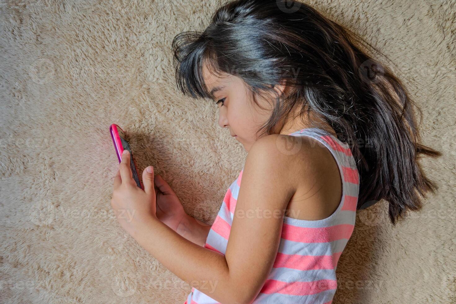 Cute little girl using a smartphone. Kid playing with mobile phone, lying on a bed. Freetime. Technology and internet concept. Toddler laying on bed and looking at smartphone. photo