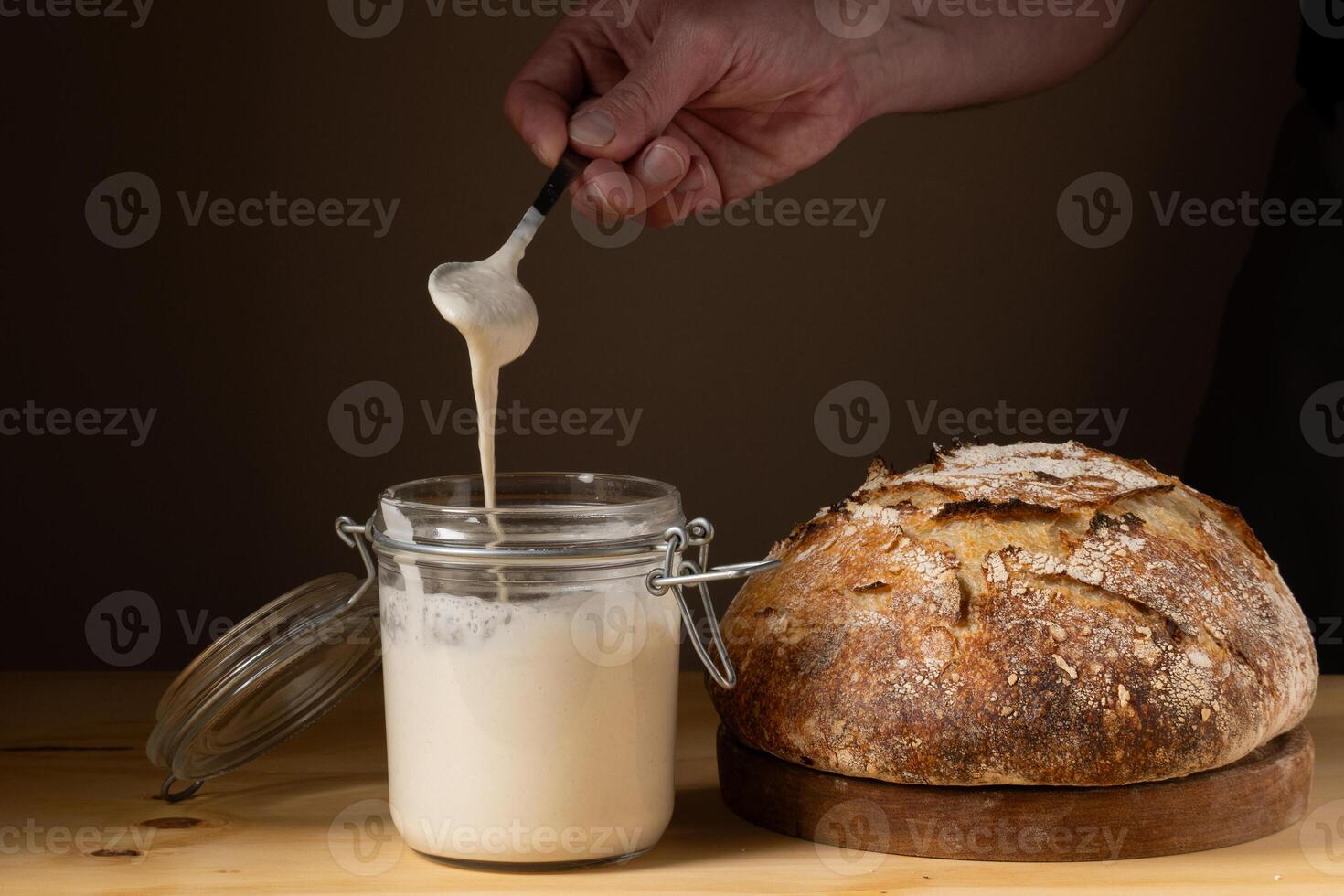 The hands of a young man showing what the sourdough is like in a glass jar. With a crusty sourdough bread next to him. photo