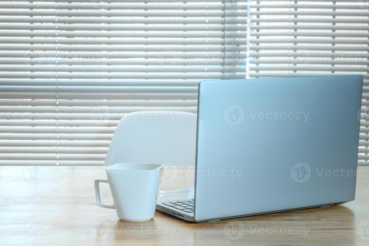 ordenador portátil y taza en el mesa en el Mañana con un antecedentes de americano persianas foto