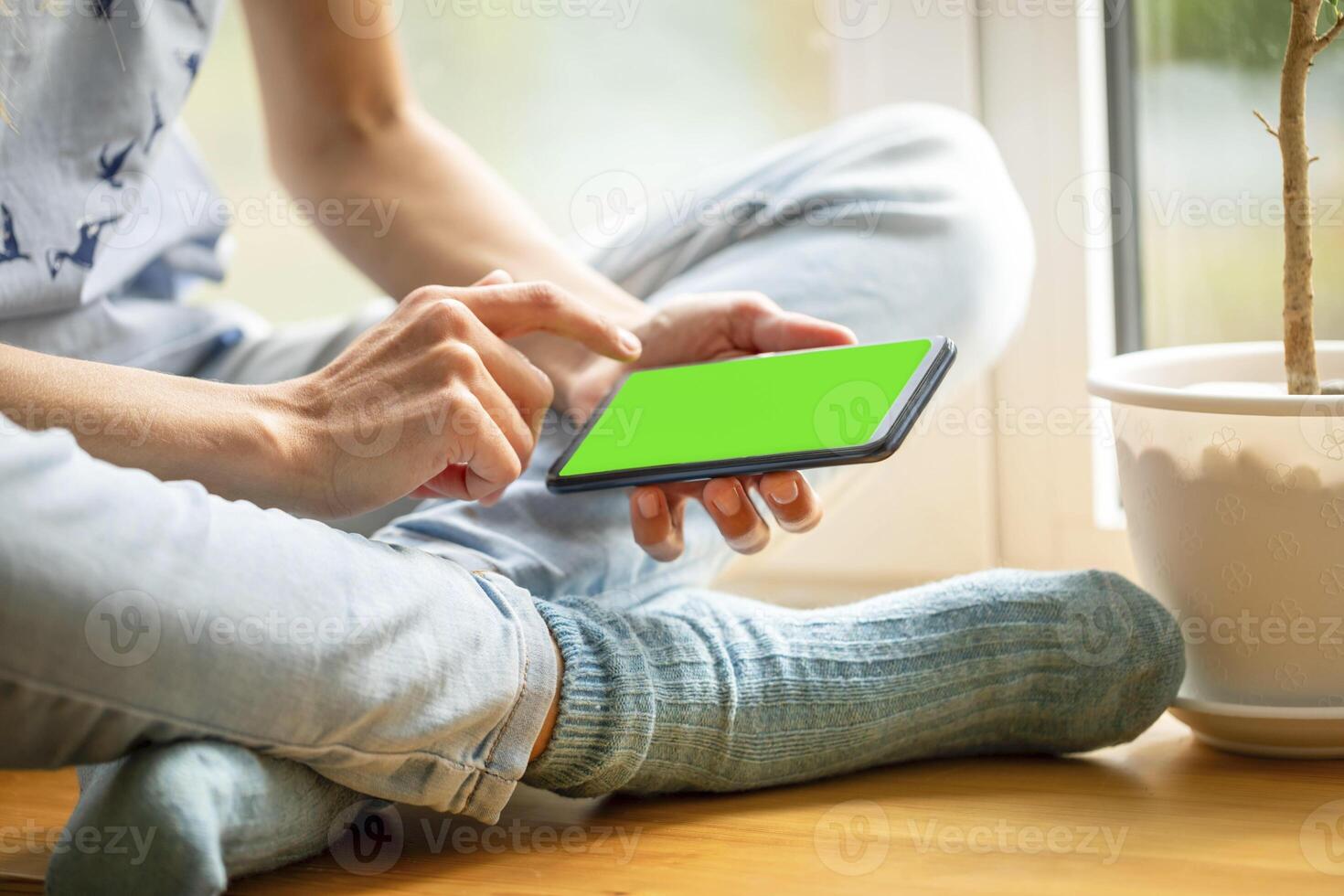 Young girl in blue jeans sitting on window kill with smartphone with green screen clicking on screen photo