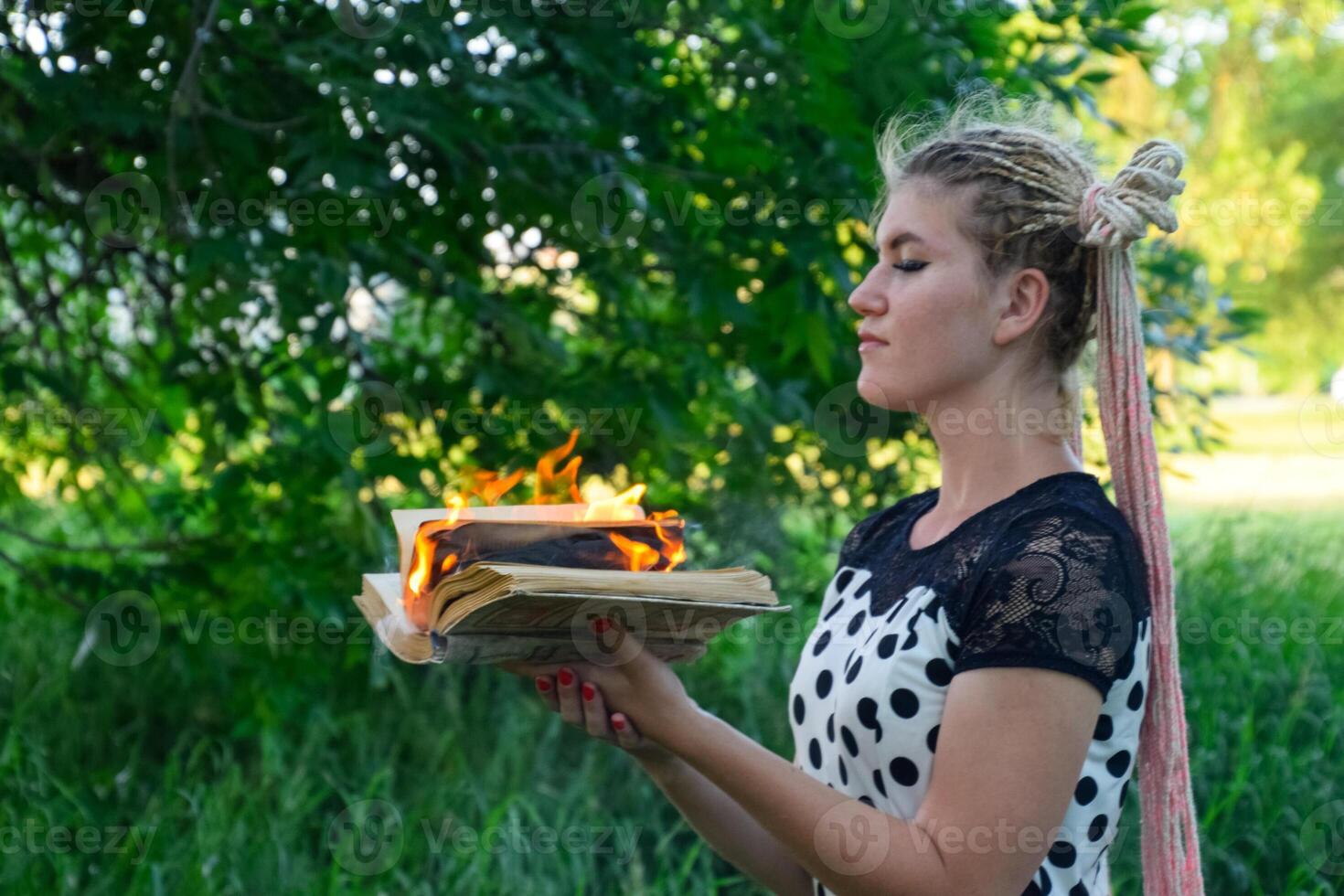 girl holds a burning book in her hands. A young woman in a forest burns a book. photo