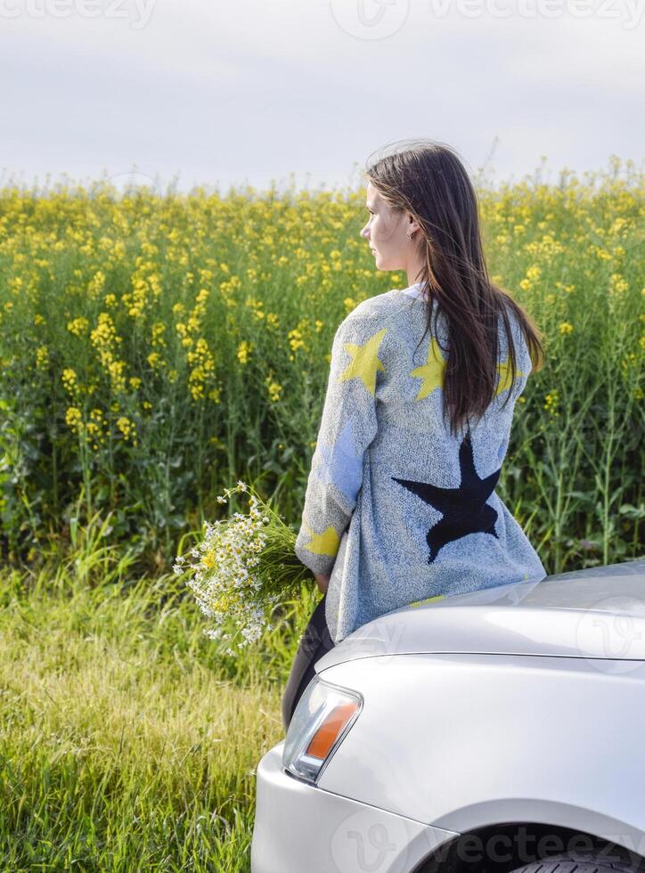 un niña con un ramo de flores de margaritas se sienta en el capucha de el coche y mira dentro el distancia. foto