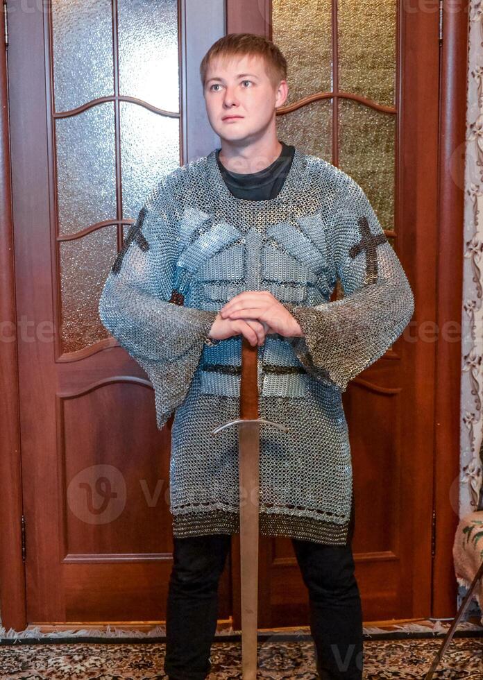 A man dressed up in a knight's hauberk at home in his room. Knight's sword in the hands of a man. photo
