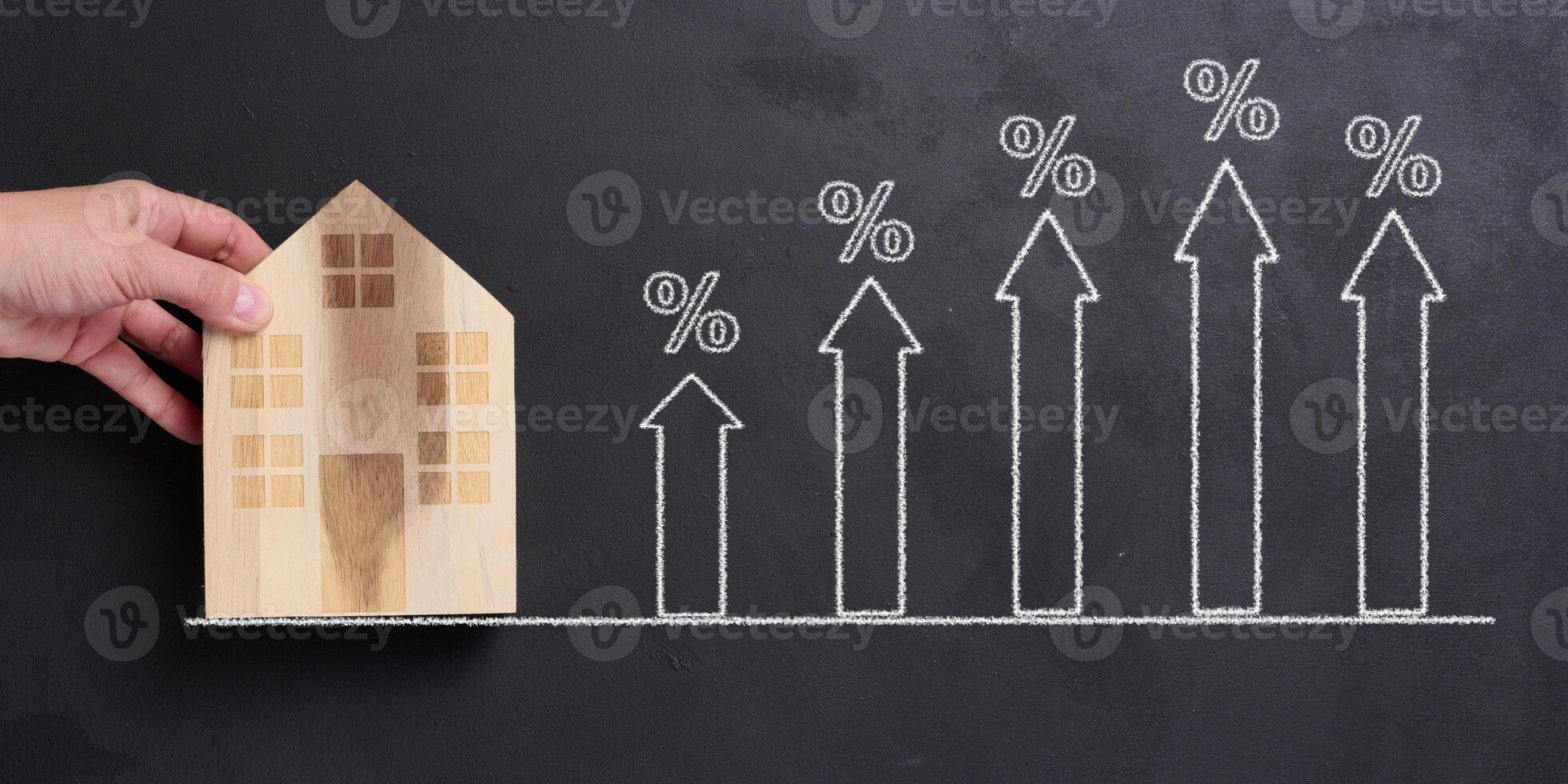 A woman's hand holds a miniature wooden house, and nearby there are drawn upward arrows with percentage signs photo