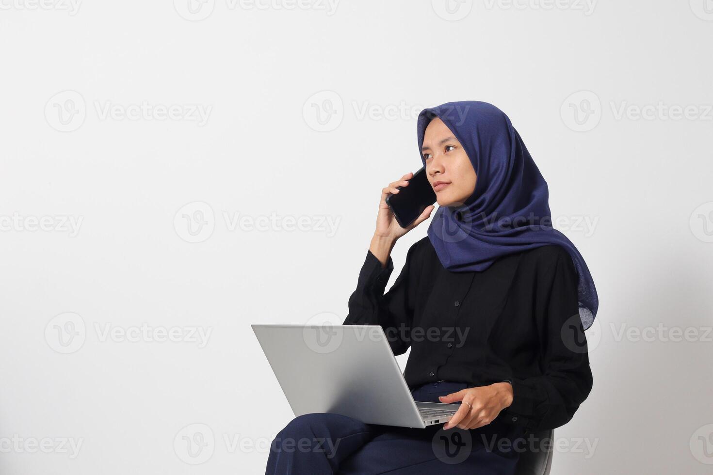Portrait of excited Asian hijab woman in casual shirt sitting on chair, working on laptop while having conversation on smartphone call. Businesswoman concept. Isolated image on white background photo