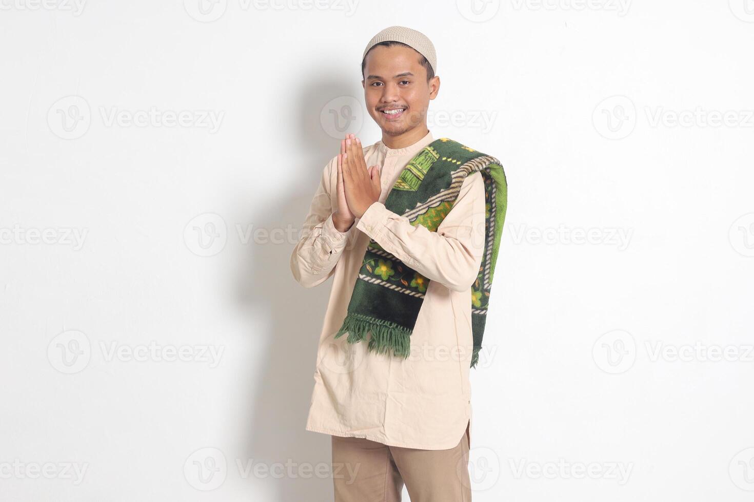 Portrait of attractive Asian muslim man in koko shirt with prayer mat showing apologize and welcome hand gesture. Apology during eid mubarak. Isolated image on white background photo