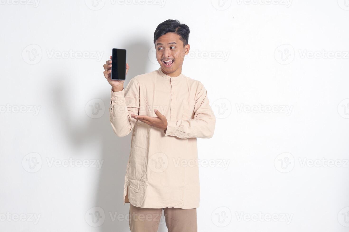 retrato de joven emocionado asiático musulmán hombre en koko camisa demostración blanco pantalla móvil teléfono Bosquejo mientras señalando y presentación producto. social medios de comunicación concepto. aislado imagen en blanco antecedentes foto