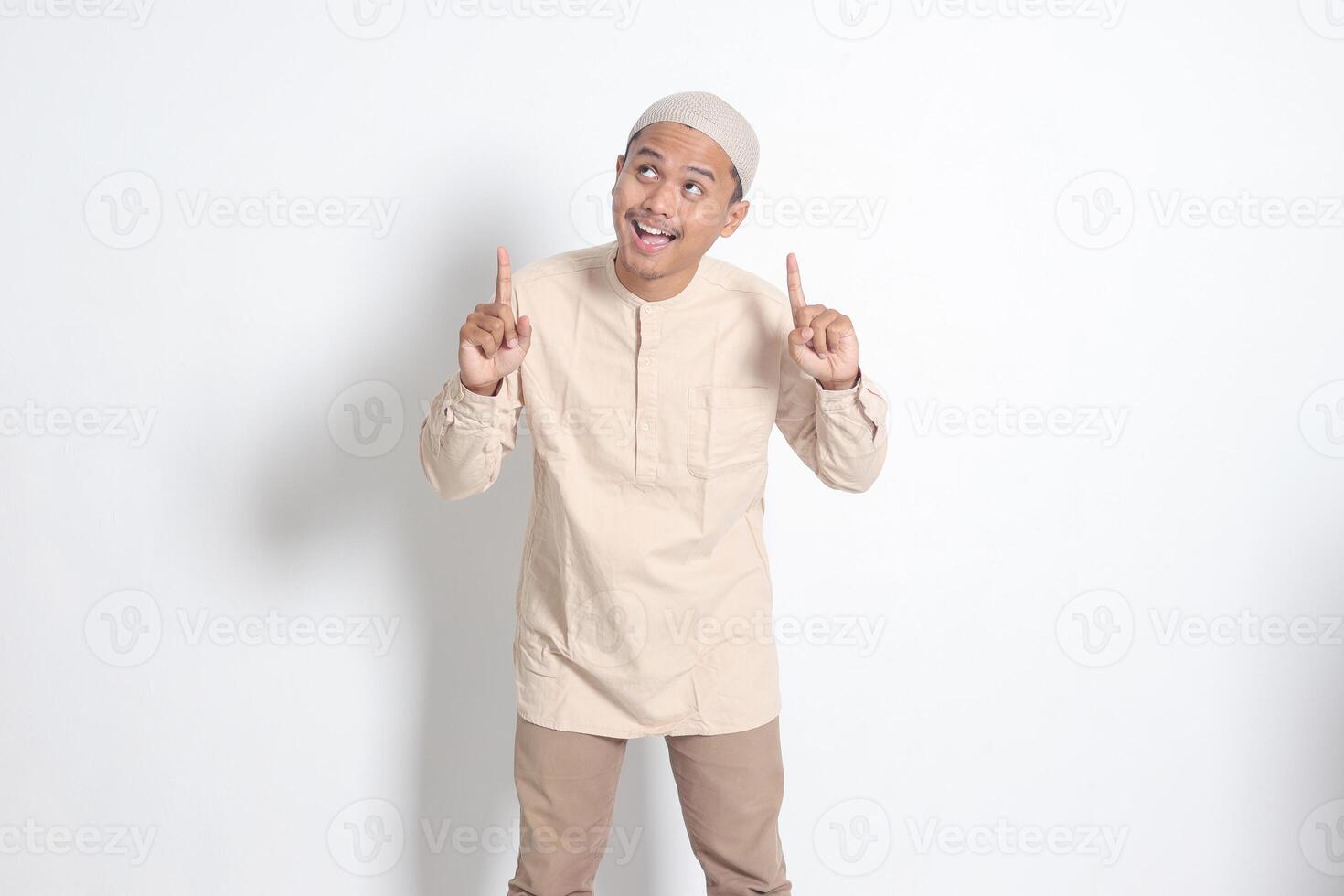 retrato de conmocionado asiático musulmán hombre en koko camisa con casquete demostración producto y señalando con su mano y dedo arriba. publicidad concepto. aislado imagen en blanco antecedentes foto