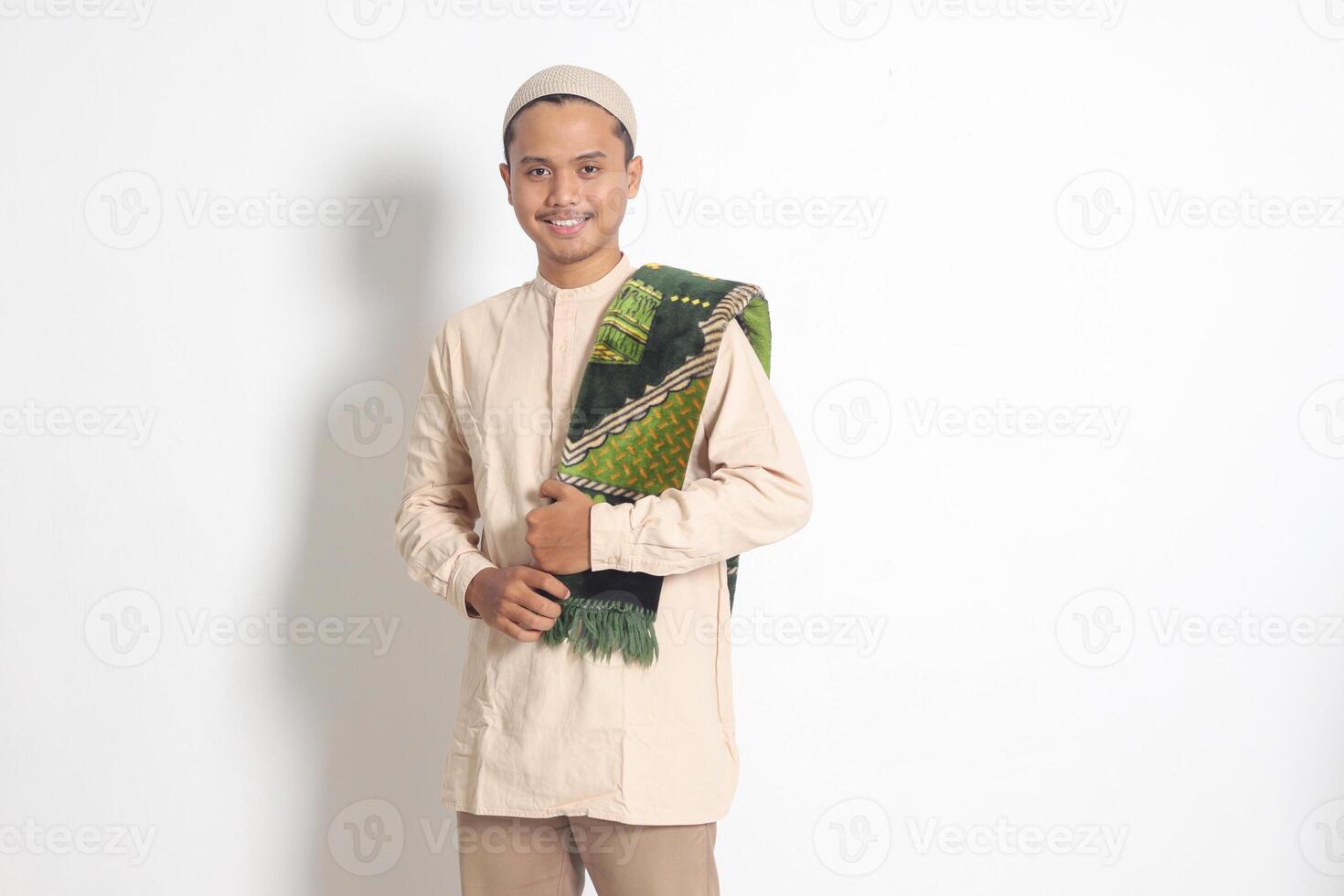 Portrait of religious Asian muslim man in koko shirt with prayer mat standing with cool pose and holding his sajadah. Isolated image on white background photo