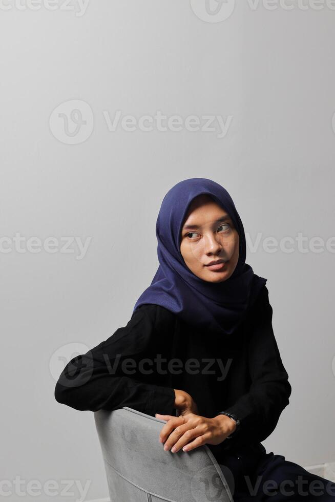 Serious Asian hijab woman in casual shirt sitting on chair, posing with various hand gesture. Isolated studio portrait on gray background. photo