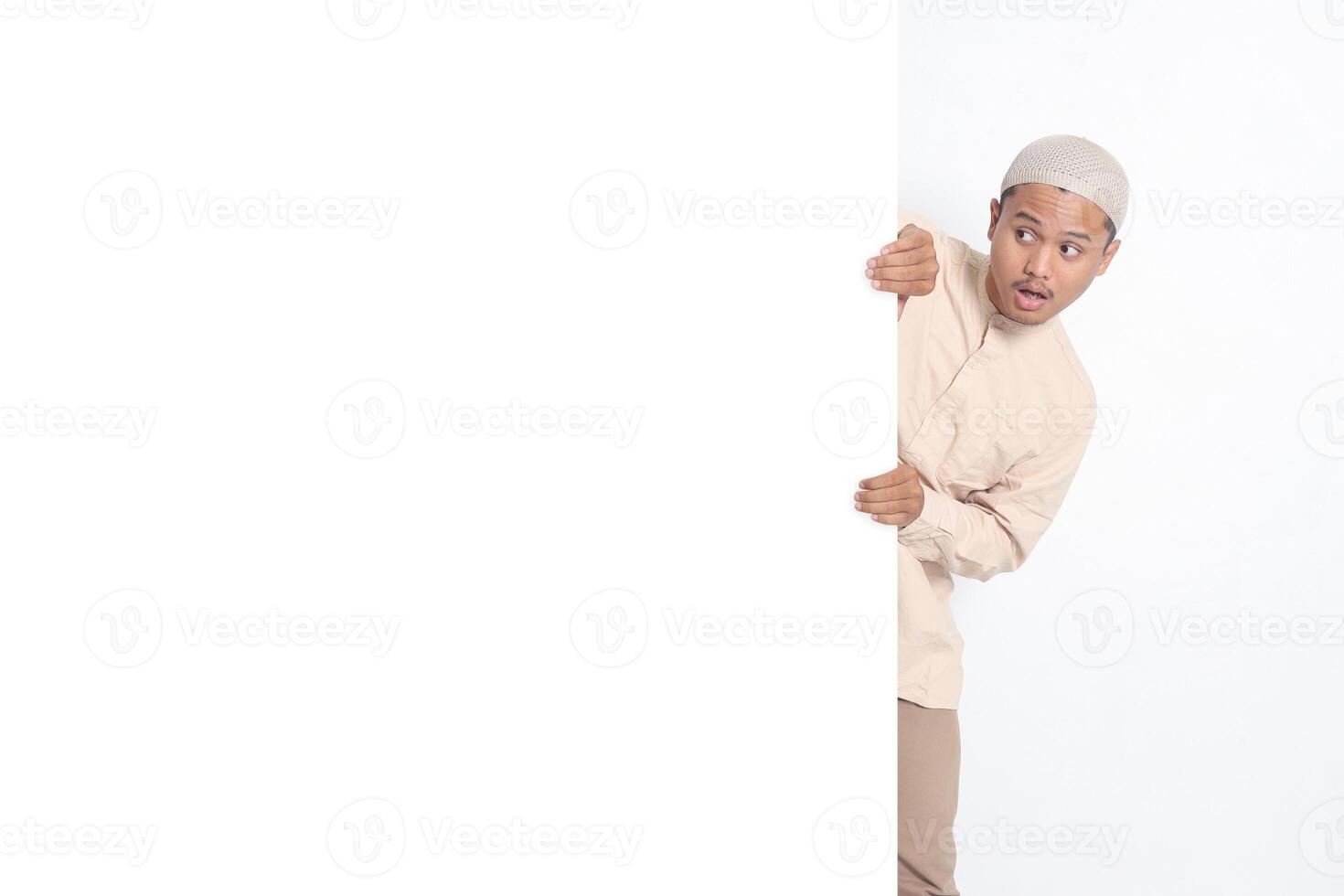 Portrait of excited Asian muslim man in koko shirt hiding behind empty white billboard with mockup template and copy space for graphic design. Advertising poster. Isolated image on white background photo