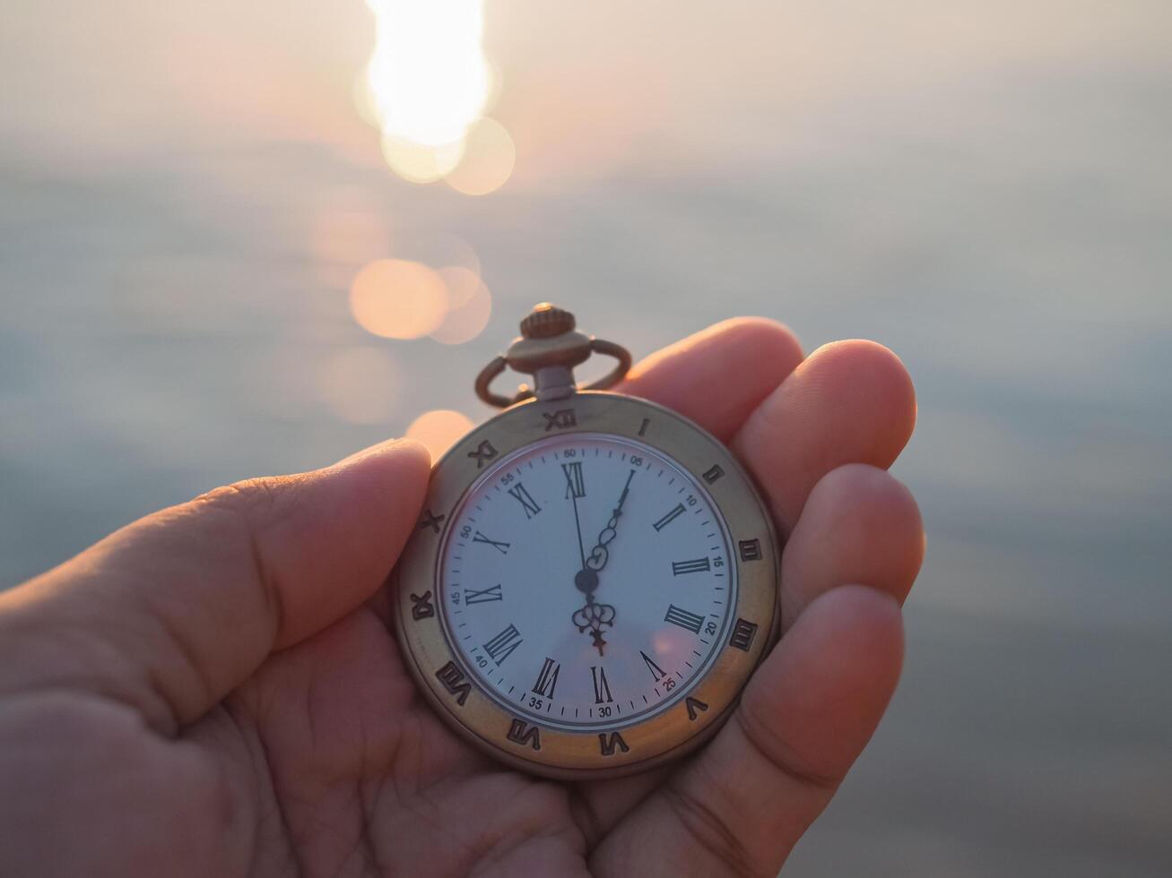 cerca arriba de Clásico bolsillo reloj en mujer mano con mar en el puesta de sol hora como antecedentes. foto