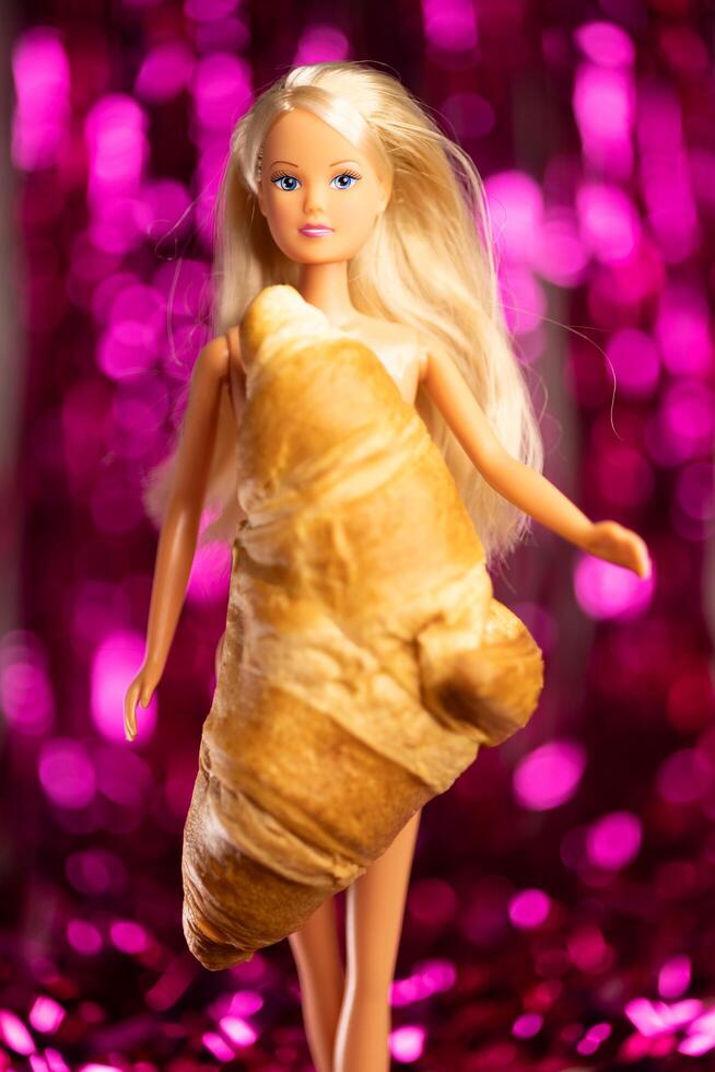 Kyiv, Ukraine, December 16, 2023, Plastic Barbie blond doll dressed in croissant instead of dress on pink foil fringe curtains background photo
